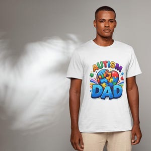 May include: White t-shirt with a colorful graphic that reads "AUTISM DAD". The design includes a heart-shaped puzzle piece and a fist. The shirt is worn by a person in a studio setting.