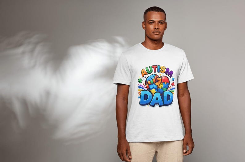 May include: White t-shirt with a colorful graphic that reads "AUTISM DAD". The design includes a heart-shaped puzzle piece and a fist. The shirt is worn by a person in a studio setting.