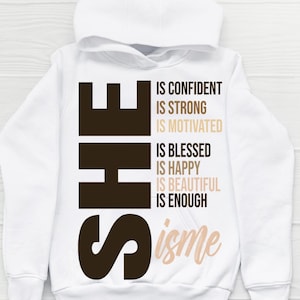 May include: White hooded sweatshirt with the word "SHE" in dark brown block letters. The text "IS CONFIDENT, IS STRONG, IS MOTIVATED, IS BLESSED, IS HAPPY, IS BEAUTIFUL, IS ENOUGH" is printed in various shades of brown. The word "isme" is written in a cursive font.
