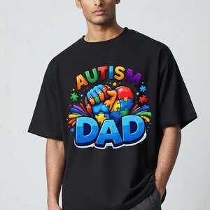 May include: Black t-shirt with a colorful graphic that reads "AUTISM DAD." The design features a heart-shaped puzzle with a fist holding it, surrounded by puzzle pieces and rainbow accents. The shirt is worn by a person.