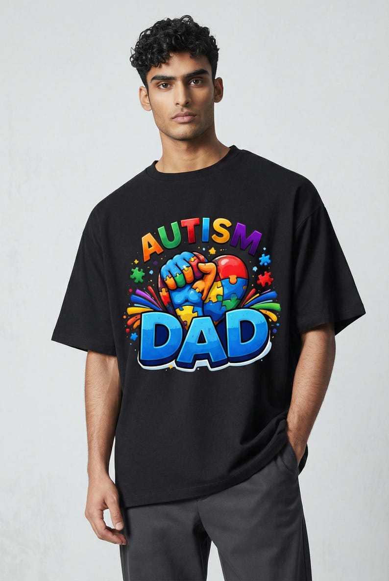 May include: Black t-shirt with a colorful graphic that reads "AUTISM DAD." The design features a heart-shaped puzzle with a fist holding it, surrounded by puzzle pieces and rainbow accents. The shirt is worn by a person.
