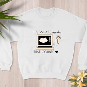 Puede incluir: Sudadera blanca con un gráfico negro de una imagen de ultrasonido y una varita. El texto dice "Es lo que está dentro lo que cuenta".