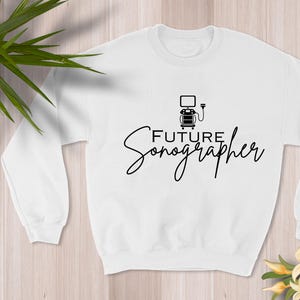 Puede incluir: Sudadera blanca con la frase "Future Sonographer" en fuente negra. El diseño incluye un pequeño gráfico de un ecógrafo. La sudadera es de cuello redondo.