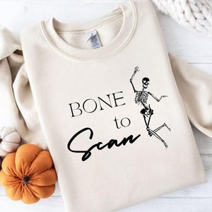 以下が含まれることがあります： 「BONE to Scan」の文字と黒い骸骨のグラフィックが特徴のベージュのスウェットシャツ。スウェットシャツは装飾的なカボチャに囲まれています。ハロウィーンのフェスティバルデザイン。