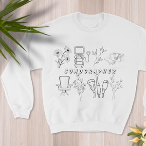 May include: White sweatshirt featuring a black line drawing of sonography equipment, flowers, and the word "SONOGRAPHER". The design includes a sonogram machine, chair, and other medical tools. The sweatshirt is a crew neck style.