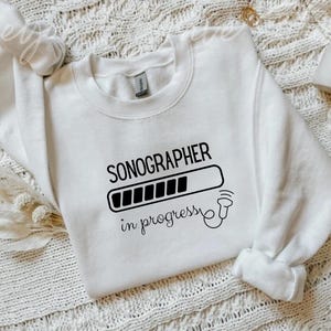 Może przedstawiać: Biała bluza z okrągłym dekoltem z czarnym napisem "SONOGRAPHER in progress". Projekt zawiera pasek ładowania i obraz USG. Bluza jest prezentowana na teksturowanej, kremowej powierzchni.