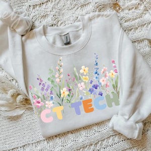 May include: A white sweatshirt with a floral design and the text "CT TECH". The flowers are in pastel shades of pink, purple, yellow, and blue. The sweatshirt is made of a soft material.