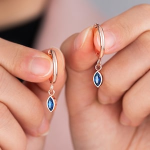 Puede incluir: Pendientes de aro de oro rosa con una gema azul oscuro en forma de marquesa colgante. Los pendientes se muestran sobre un fondo rosa.