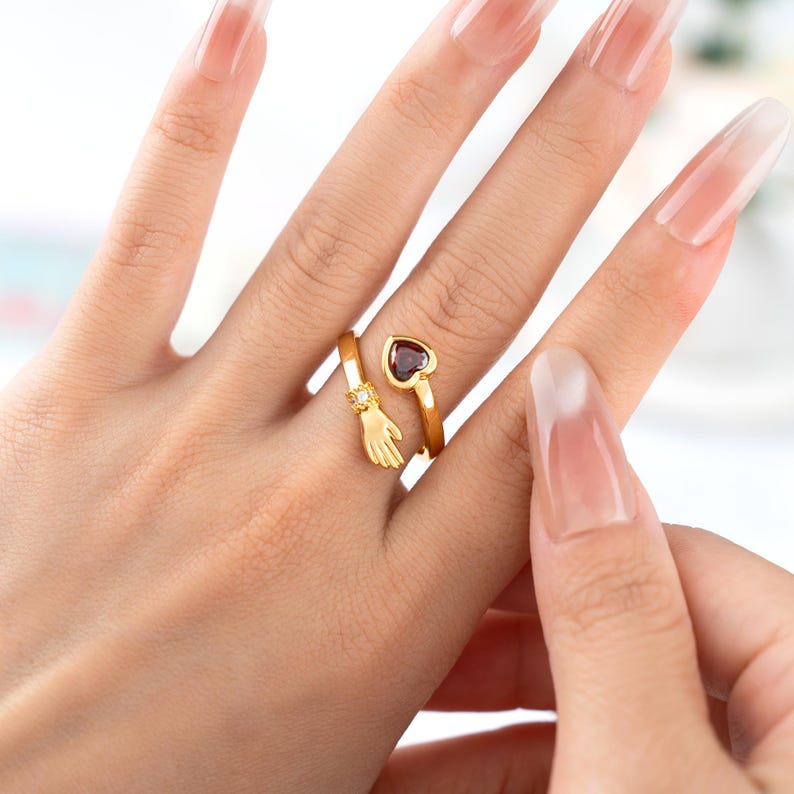 K&ouml;nnte beinhalten: Ein goldfarbener Ring mit einem herzf&ouml;rmigen, dunkelroten Edelstein und einem kleinen Handdesign. Der Ring umschlie&szlig;t den Finger, wobei die Hand das Band umfasst. Ein kleiner Diamant befindet sich auf der Hand.