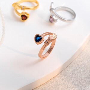 K&ouml;nnte beinhalten: Ros&eacute;goldener Ring mit einem herzf&ouml;rmigen blauen Edelstein, der von einem Handdesign gehalten wird. Auf dem Ring ist das Wort "Kulana" eingraviert. Weitere Ringe in Gold und Silber mit herzf&ouml;rmigen Edelsteinen sind ebenfalls sichtbar.