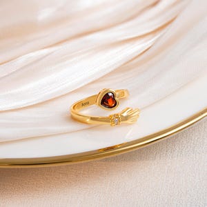K&ouml;nnte beinhalten: Ein goldfarbener Ring mit einem herzf&ouml;rmigen, roten Edelstein und einem kleinen Diamanten. Der Ring hat ein Design mit umarmender Hand und den Namen "Anna" auf dem Band eingraviert. Der Ring wird auf einem wei&szlig;en Teller mit Goldrand pr&auml;sentiert.