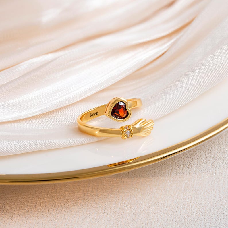 K&ouml;nnte beinhalten: Ein goldfarbener Ring mit einem herzf&ouml;rmigen, roten Edelstein und einem kleinen Diamanten. Der Ring hat ein Design mit umarmender Hand und den Namen "Anna" auf dem Band eingraviert. Der Ring wird auf einem wei&szlig;en Teller mit Goldrand pr&auml;sentiert.