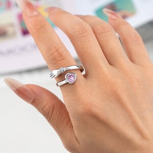K&ouml;nnte beinhalten: Ein silberfarbener Ring mit einem Handdesign, das einen rosa herzf&ouml;rmigen Stein umarmt. Die Hand ist mit einem kleinen, klaren Edelstein verziert. Der Ring wird an einem Finger getragen und zeigt sein zartes Design und romantisches Thema.