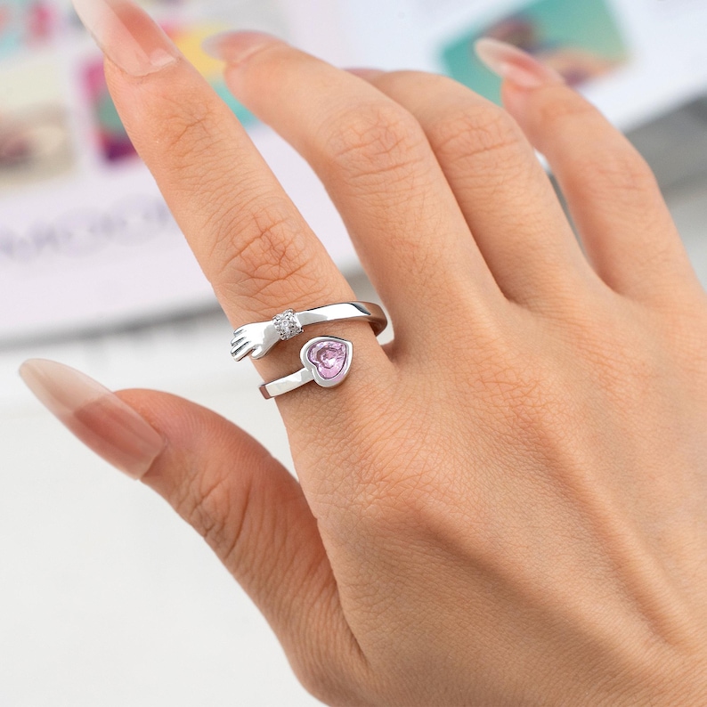 K&ouml;nnte beinhalten: Ein silberfarbener Ring mit einem Handdesign, das einen rosa herzf&ouml;rmigen Stein umarmt. Die Hand ist mit einem kleinen, klaren Edelstein verziert. Der Ring wird an einem Finger getragen und zeigt sein zartes Design und romantisches Thema.