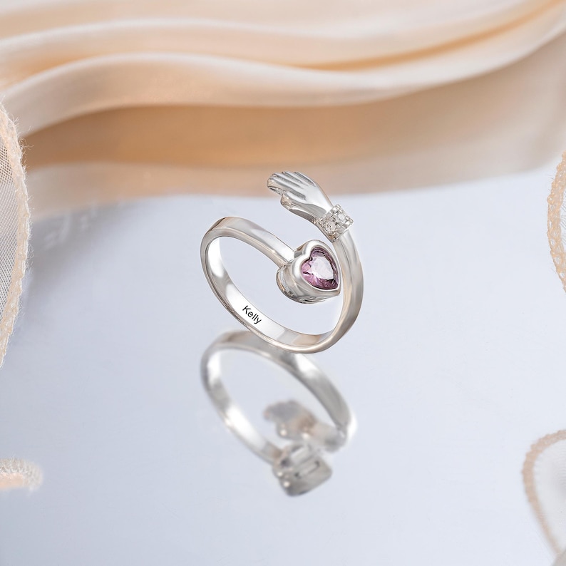 K&ouml;nnte beinhalten: Ein silberner Ring mit einem herzf&ouml;rmigen rosa Edelstein und einem Handmotiv. Der Ring tr&auml;gt den eingravierten Namen "Kelly". Das Handmotiv ist mit kleinen, klaren Edelsteinen verziert.