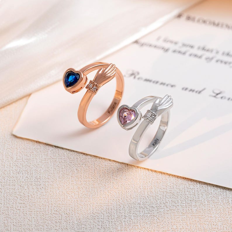 K&ouml;nnte beinhalten: Zwei offene Handringe, einer in Ros&eacute;gold und einer in Silber, die jeweils einen herzf&ouml;rmigen Edelstein halten. Der Ros&eacute;goldring hat einen blauen Edelstein, und der Silberring hat einen rosa Edelstein.
