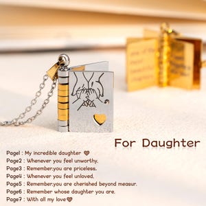 Peut inclure: Un collier avec un médaillon en forme de livre, argenté et doré. La couverture avant présente un dessin de mains et un cœur. Le texte "For Daughter" est visible. Le médaillon s'ouvre sur des pages dorées avec du texte.