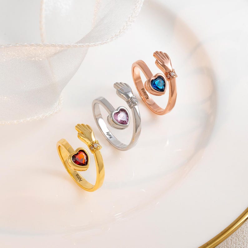 K&ouml;nnte beinhalten: Drei offene Ringe mit herzf&ouml;rmigen Edelsteinen in Rot, Lila und Blau. Jeder Ring hat ein Handdesign auf beiden Seiten des Herzens. Die Ringe sind gold-, silber- und ros&eacute;goldfarben.