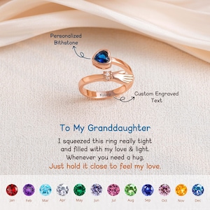 Könnte beinhalten: Roségoldener Ring mit herzförmigem blauem Edelstein und kleinem Diamantakzent. Der Ring hat ein Umarmungsdesign mit eingraviertem Text. Der Text auf dem Ring lautet "Kulana". Der Ring ist ein Geschenk für eine Enkelin.
