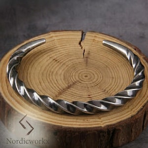 Könnte beinhalten: Ein silberfarbenes, verdrehtes Metallarmband. Das Armband hat ein spiralförmiges Design und ist an den Enden offen. Das Armband wird auf einer Holzoberfläche mit sichtbarer Maserung und einem Riss präsentiert. Der Markenname "Nordicworkx" ist sichtbar.