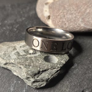 May include: A silver-toned metal ring with a brushed finish featuring the engraved text ONE LO visible on the outer band. The ring has a flat profile and a smooth interior surface, showcasing a clean and simple design with clear lettering.
