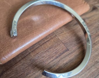 May include: A silver-coloured, open cuff bracelet. The bracelet has a smooth, rounded design and is resting on a brown leather surface. The bracelet is made of a shiny metal and has a simple, minimalist style.