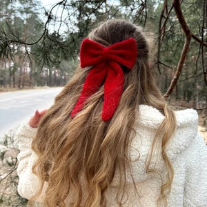 以下が含まれることがあります： 赤いリボンのヘアアクセサリーが、この画像の中心です。リボンは長く、ウェーブのかかったブロンドの髪に付けられています。人物は白くふわふわしたジャケットを着ています。背景は、木々のあるぼやけた屋外のシーンです。