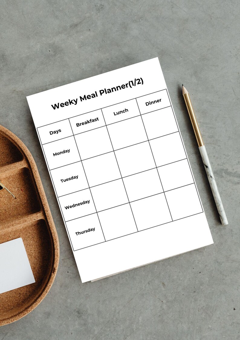 May include: A white weekly meal planner with the title "Weeky Meal Planner (1/2)" and a grid for daily meal planning. A gold and marble pen rests on the right side. A wooden tray is on the left side.
