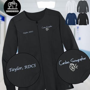 Könnte beinhalten: Schwarze medizinische Scrub-Jacke mit langen Ärmeln und Reißverschluss. Die Jacke hat gestickten Text mit der Aufschrift "Taylor, RDCS" und "Cardiac Sonographer" mit einer Herzgrafik. Zusätzliche Farboptionen sind in Weiß, Grau, Marineblau und Schwarz abgebildet.