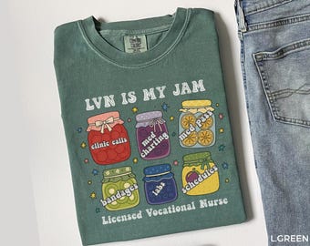 以下が含まれることがあります： 「LVN IS MY JAM」の文字と、「clinic calls」、「med charting」、「med pass」、「bandages」、「labs」、「schedules」とラベル付けされた瓶のイラストが描かれた緑色のTシャツ。「Licensed Vocational Nurse」の文字が下にあります。