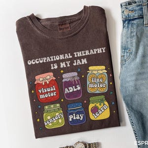 Może przedstawiać: Brązowa koszulka z napisem "OCCUPATIONAL THERAPHY IS MY JAM" i kolorowymi ilustracjami słoików. Słoiki zawierają słowa "visual motor", "ADLS", "fine motor", "sensory", "play" i "learn". Widoczne są jasnoniebieskie dżinsy.
