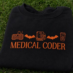 Puede incluir: Una sudadera negra de cuello redondo con bordado naranja. El diseño incluye un teclado, un murciélago, un libro médico CPT con una lupa, otro murciélago y un vaso de café. El texto MEDICAL CODER está bordado en color naranja debajo.