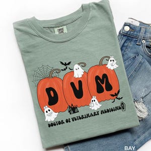 May include: A sage green t-shirt with a Halloween-themed design. Three orange pumpkins spell out "DVM" with ghosts, bats, and a spiderweb. The text "DOCTOR OF VETERINARY MEDICINE" is below the pumpkins. Denim shorts are next to the shirt.