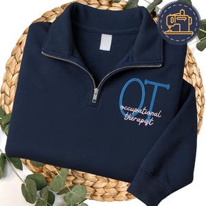May include: Navy blue quarter-zip sweatshirt with the embroidered letters "OT" in blue, and the words "occupational therapist" in pink. The sweatshirt has a white tag on the collar and a silver zipper. The sweatshirt is on a woven basket.