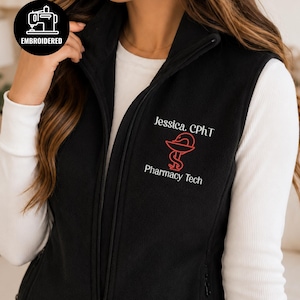 Puede incluir: Un chaleco de forro polar negro con cremallera frontal completa y cuello alto. El pecho izquierdo presenta texto bordado en blanco que dice Jessica. CPhT y Pharmacy Tech, con un símbolo rojo bordado de la copa de Higía en el centro.