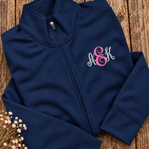 May include: A navy blue zip-up jacket featuring a high collar and a central zipper closure. The left chest area displays an embroidered monogram with the letters A, E, and K in a cursive font, with the central E in pink and A and K in white.
