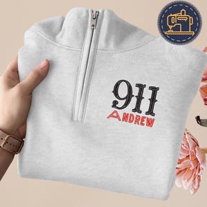 May include: A light gray quarter-zip sweatshirt with the embroidered text "911 ANDREW" in black and red. The sweatshirt has a silver zipper and a small patch with a sewing machine design.
