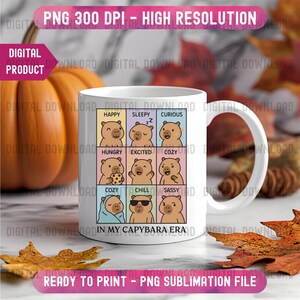 May include: White ceramic mug featuring a grid of capybara illustrations expressing different emotions, with text that reads "IN MY CAPYBARA ERA." The mug is surrounded by autumn leaves and a pumpkin.