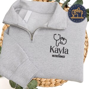 May include: A pale grey quarter-zip sweatshirt featuring a black stethoscope and apple design, with the name "Kayla" and the word "Nutritionist" printed below. The sweatshirt is folded on a woven basket.