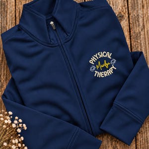May include: A navy blue zip-up jacket featuring a mock neck collar and a central zipper. Embroidered on the left chest is the text "PHYSICAL THERAPY" in a circular arrangement with the name "Marilyn" in yellow script in the center.
