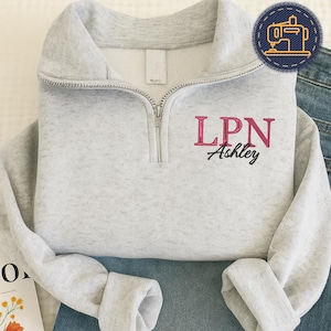 Può includere: Una felpa grigio chiaro con scollo a zip. La felpa presenta la scritta ricamata "LPN Ashley" in rosa e nero. Le maniche sono a polsino e il capo è appoggiato su denim blu.