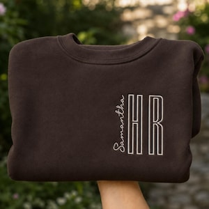 Peut inclure: Un sweat-shirt à col rond marron foncé avec une broderie blanche sur le devant. La broderie présente le prénom Samantha en écriture cursive positionné verticalement à côté des initiales HR en grandes lettres capitales au style épuré.