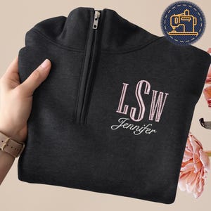 May include: A dark gray quarter-zip sweatshirt with a light pink embroidered monogram and name. The sweatshirt has a silver zipper and is folded. The monogram reads "LSW" above the name "Jennifer".