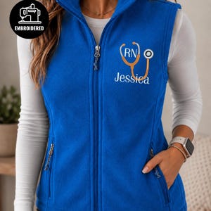May include: A royal blue fleece vest featuring a full-length front zipper, two zippered side pockets, and a stand-up collar. The chest is embroidered with an orange stethoscope, the letters RN, and the name Jessica in white script lettering.