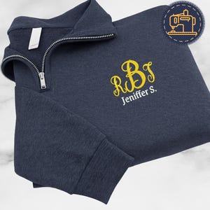 Może przedstawiać: Granatowa bluza z zamkiem błyskawicznym do połowy szyi ze złotym monogramem liter "RBS" i imieniem "Jeniffer S." wyhaftowanym z przodu. Bluza ma zapięcie na zamek i małą metkę na kołnierzu.