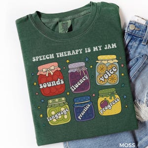 Könnte beinhalten: Moosgrünes T-Shirt mit dem Text "SPEECH THERAPY IS MY JAM". Das Shirt zeigt Illustrationen von sechs Marmeladengläsern, die jeweils mit Begriffen der Sprachtherapie beschriftet sind: Sounds, Fluency, Voice, Language, Practice und Progress. Das Shirt wird mit Jeansshorts gezeigt.
