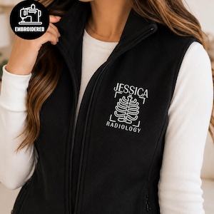 May include: A black fleece vest featuring a full-length front zipper and a high collar. The chest area displays white embroidery with the text JESSICA RADIOLOGY and a stylized rib cage graphic. A circular black badge reads EMBROIDERED.
