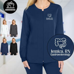 Può includere: Giacca da medico blu navy ricamata con chiusura a zip e due tasche anteriori. La giacca presenta la scritta "Jessica, RN Gastroenterology" e un logo ricamato bianco. Sono mostrate opzioni di colore aggiuntive.