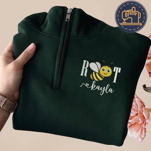 May include: A dark green quarter-zip sweatshirt with a bee and the name "kayla" embroidered on the front. The bee is yellow and black with a smiling face. The sweatshirt has a zipper and a small embroidered patch.