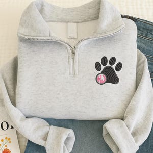 Puede incluir: Sudadera gris claro con cremallera de cuarto de cremallera, diseño de huella de pata negra y monograma rosa. La sudadera está doblada, con las mangas y el dobladillo inferior visibles. La cremallera es plateada.